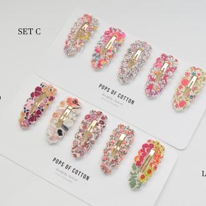 May include: Two sets of hair clips with floral patterns. The top set is labeled "SET C" and the bottom set is labeled "SET D". Both sets are made with Liberty Fabric and are sold by Pops of Cotton.
