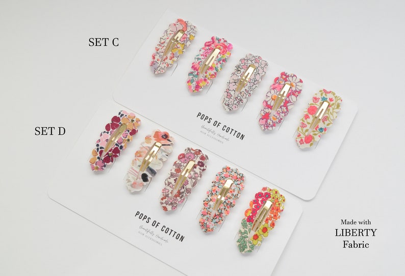 May include: Two sets of hair clips with floral patterns. The top set is labeled "SET C" and the bottom set is labeled "SET D". Both sets are made with Liberty Fabric and are sold by Pops of Cotton.