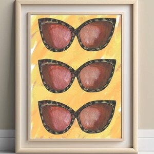 May include: A framed print showing three pairs of cat-eye sunglasses. The sunglasses have tortoiseshell frames and reddish-brown lenses. The background is a vibrant yellow with brushstroke textures. The artwork is displayed in a light-coloured wooden frame.