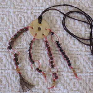May include: A wooden bead necklace with a round wooden pendant and brown leather cord. The necklace has five strands of beads, each with a different color and pattern. The beads are brown, red, and black.