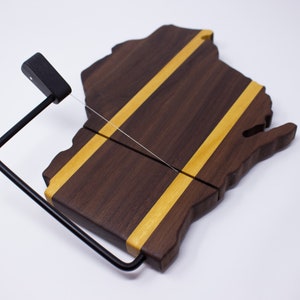 Könnte beinhalten: Ein Käseboard aus Holz in Form des Bundesstaates Wisconsin mit zwei gelben Streifen. Ein schwarzer Käse-Drahtcutter ist an der Platte befestigt.
