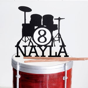 May include: A red and silver drum cake decorated with a black silhouette of a drum set and the name "Nayla" and the number "8".