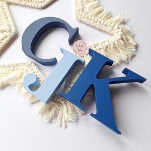 May include: Three dimensional letters C, J, and K in varying shades of blue. The letter C is dark blue, the J is light blue, and the K is a medium blue. A small circular sticker with the text Perfecto Arte is placed between the letters.