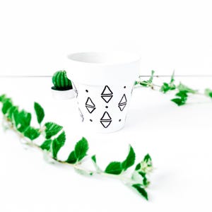 May include: A white ceramic plant pot with a geometric black diamond pattern. The pot is surrounded by green foliage and a small green cactus. The design is minimalist and modern, suitable for indoor plants.
