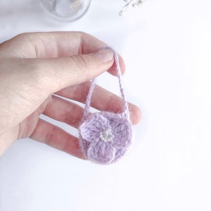 May include: A small, lavender crocheted purse with a flower design and a pearl center. The purse has a matching handle and is held in a hand against a white background. The purse is approximately 2 inches in diameter.