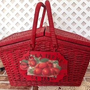 May include: A bright red woven wicker picnic basket with two hinged lids and two upright handles. A rectangular sign hangs from the front, featuring a graphic of red peaches and the text DE LA FERME A LA TABLE in black lettering.