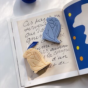 May include: Two small, decorative sculptures of figures in a seated pose. The sculptures are made of a smooth, light-colored material. The figure on the left is a light blue color, and the figure on the right is a light beige color. The sculptures are sitting on a page of a book with handwritten text.