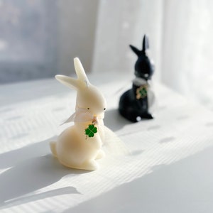 May include: Two white and black rabbit shaped candles with a green four-leaf clover charm. The white rabbit candle has a white ribbon around its neck.