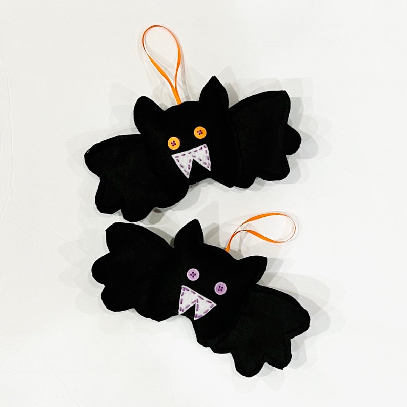May include: Two black felt bat ornaments with orange ribbon hangers. Each bat has button eyes and a stitched white mouth with purple stitching. The bats are a Halloween decoration.