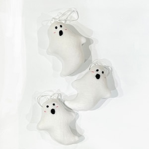May include: Three white felt ghost ornaments with black eyes and open mouths, and pink blush. Each ghost has a white string for hanging. The ornaments are arranged on a white background.