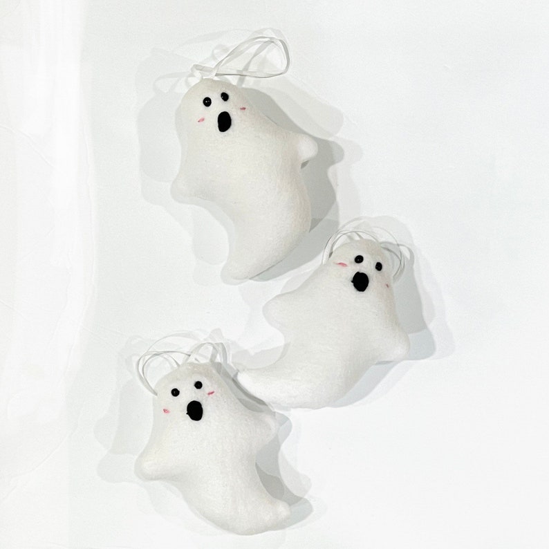 May include: Three white felt ghost ornaments with black eyes and open mouths, and pink blush. Each ghost has a white string for hanging. The ornaments are arranged on a white background.