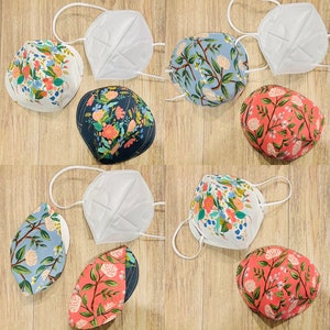 May include: Assortment of face masks with floral patterns and white KN95 masks. The floral masks feature designs in various colors, including pink, blue, and green. The masks are arranged on a light brown wooden surface.