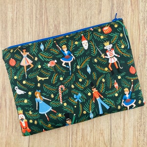 May include: A rectangular, zippered pouch with a festive Christmas-themed pattern. The dark green fabric features illustrations of ballerinas, nutcrackers, and ornaments. The zipper is blue, and the pouch is likely for cosmetics or small items.