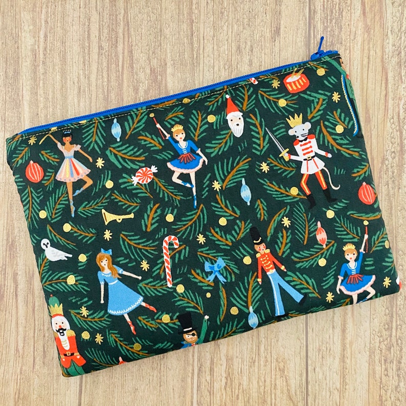 May include: A rectangular, zippered pouch with a festive Christmas-themed pattern. The dark green fabric features illustrations of ballerinas, nutcrackers, and ornaments. The zipper is blue, and the pouch is likely for cosmetics or small items.
