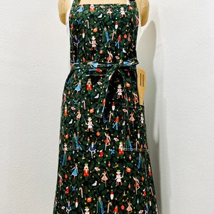 May include: A dark green apron with a festive Nutcracker print. The apron features a repeating pattern of Nutcracker figures, ballerinas, and candy canes. It has a waist tie, two front pockets, and a wooden spatula in the right pocket.