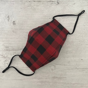 May include: A red and black plaid face mask with adjustable black ear loops. The mask is made of fabric and has a classic buffalo check pattern. The mask is designed to cover the nose and mouth.