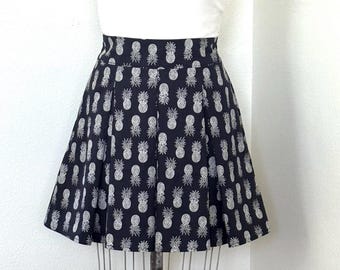 Falda corta de algodón negro con estampado de piña hawaiana, plisada en la parte delantera con bolsillos laterales y fruncido elástico en la parte trasera.