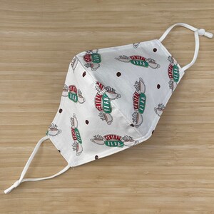 May include: White face mask with a repeating pattern of the "Central Perk" logo from the TV show "Friends" and brown polka dots. The mask has adjustable white ear loops.