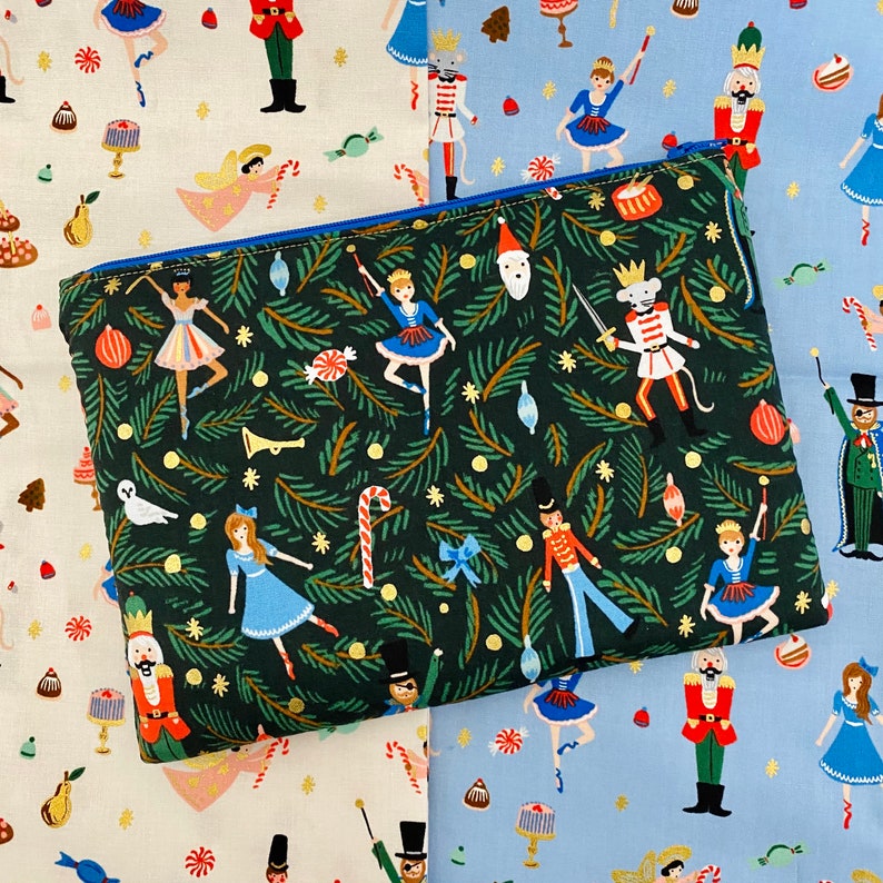 May include: A rectangular, zippered pouch with a dark green background featuring a repeating pattern of Christmas-themed illustrations. The illustrations include ballerinas, nutcrackers, and candy canes. The zipper is blue.