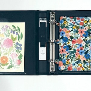 May include: Open navy blue binder with a floral print pouch and a floral print card. The pouch is filled with colorful pens. The card features a variety of colorful flowers. The binder has a metal ring mechanism.