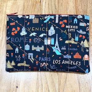 May include: A black quilted travel pouch featuring illustrations of landmarks and city names like Venice, Rome, Paris, and Los Angeles. The pouch has a red zipper and a diamond-stitched pattern.