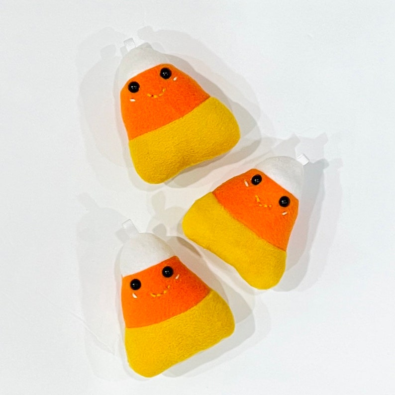 May include: Three plush candy corn ornaments with smiling faces. Each ornament features a white top, orange center, and yellow base. The ornaments are designed for Halloween or fall decor.