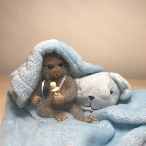 May include: A small, brown, furry toy monkey wearing a blue blanket and holding a white pacifier. The monkey is sitting on a blue and white blanket with a white teddy bear head.