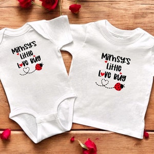 Op de afbeelding: Twee witte baby-outfits, een onesie en een t-shirt, worden gepresenteerd op een houten oppervlak. Elk kledingstuk heeft de tekst "Mimsy's Little Love Bug" in zwart schrift, met een rode lieveheersbeestje en hartafbeelding. Rode rozen omlijsten de items.
