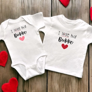 May include: Two white baby onesies with short sleeves. One onesie says "I love my Bubbe" with a pink heart. The other onesie says "I love my Bubbe" with a red heart.