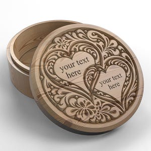 Puede incluir: Caja redonda de madera con tapa parcialmente abierta. La tapa está decorada con un diseño grabado con dos corazones y motivos florales. El texto "your text here" es visible en la tapa. La caja es de madera clara.