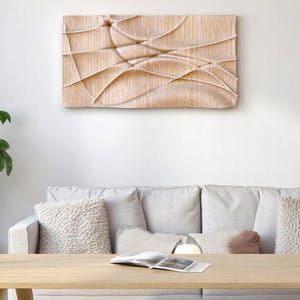 Puede incluir: Una obra de arte mural de madera de color claro con un diseño abstracto texturizado de líneas curvas. La obra de arte cuelga sobre un sofá de color claro con cojines decorativos. Una mesa de centro de madera y una planta en maceta están en primer plano.