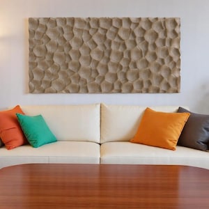 May include: A living room scene featuring a white sofa with colorful throw pillows in orange, teal, and gray. Above the sofa hangs a large, textured, beige wall art piece. A wooden coffee table sits in front of the sofa.