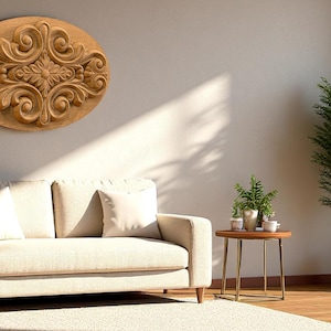 May include: A carved wooden wall decor piece with an oval shape and intricate floral and scroll design. The artwork is mounted above a beige sofa with white pillows. A small side table and a potted plant are also in the room.