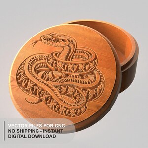 May include: A round wooden box with a lid featuring a detailed, carved relief of a coiled snake. The snake design incorporates intricate geometric patterns within its scales. The box is shown with the lid slightly offset, revealing the hollow interior.