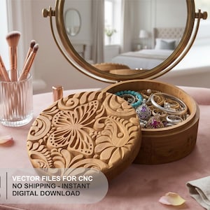 Op de afbeelding: Een ronde houten sieradendoos met een uitgesneden vlinder- en bloemenpatroon op het deksel. De open doos bevat diverse ringen en armbanden. Daarnaast staat een glazen houder met drie make-upkwasten met roségouden handvatten.