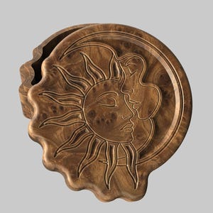 May include: A wooden box with a celestial design. The lid features a sun and moon motif, with the sun's rays radiating outwards. The box is a warm brown color, with a textured surface. The box is approximately 6 inches in diameter.