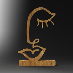 May include: Wooden sculpture of a stylised face with a closed eye, a nose, and a mouth. The sculpture is on a wooden base.