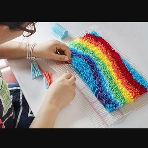 May include: A person is working on a rainbow-colored rug hooking project. The rug is made of colorful yarn and is being hooked onto a white canvas. The person is using a needle to thread the yarn through the canvas.