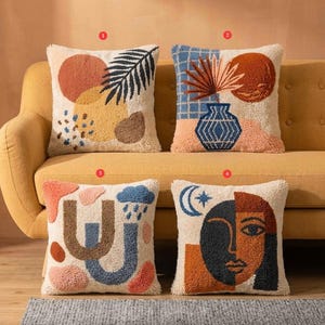 May include: Four decorative throw pillows with abstract designs. Each pillow features a cream-colored background with colorful, textured patterns. Designs include geometric shapes, botanical elements, and a face. The pillows are arranged on a yellow sofa.