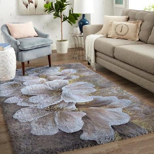 Puede incluir: Una alfombra rectangular con un diseño floral. La alfombra presenta grandes flores blancas y grises estilizadas con acentos amarillos sobre un fondo gris. La alfombra está colocada sobre un suelo de madera en un salón.
