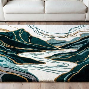 Op de afbeelding: Een rechthoekig vloerkleed met een landschapsontwerp. Het kleed toont een rivier die door turquoise en donkergroene bergen stroomt, met gouden accenten. De achtergrond is wit. Het kleed ligt op een houten vloer.