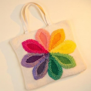 May include: A white tote bag with cream-colored handles. The bag features a large, colorful flower design in shades of red, pink, orange, yellow, green, blue, and purple. The flower is centered on the bag.