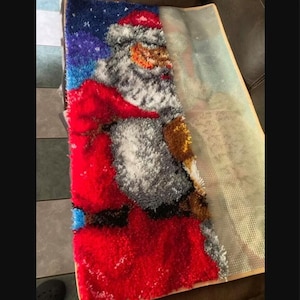 Pode incluir: Um kit de tapete de gancho parcialmente conclu&iacute;do com uma figura de Pai Natal com barba branca, casaco e gorro vermelhos sobre um fundo azul escuro com pontos brancos. O lado esquerdo mostra la&ccedil;os de fio acabados e o direito a grelha.
