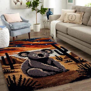 Op de afbeelding: Een rechthoekig vloerkleed met een koala beer ontwerp. Het vloerkleed heeft een woestijnlandschap met cactussen en een zonsonderganghemel in tinten oranje, blauw en bruin. De koala is grijs en zwart.