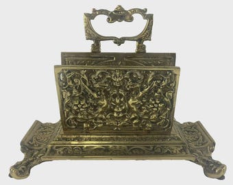 May include: Ornate brass letter holder with a handle. The letter holder features detailed relief carvings of mythical figures and floral patterns. The base has decorative feet. The overall design is antique and elegant.