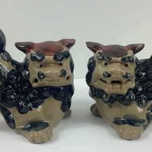 May include: A pair of ceramic guardian lion statues. Each statue has a tan body with dark blue and black accents, a red-brown head, and a large, stylized tail. The statues are in a seated position.