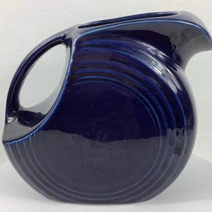 May include: A navy blue ceramic pitcher with a large handle and a curved spout. The pitcher has a glossy finish and a series of concentric, curved ridges that add texture. The design is simple and elegant, suitable for serving beverages.