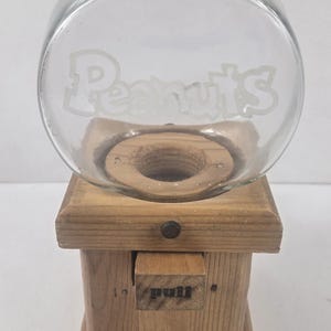 May include: A clear glass bowl with the word Peanuts etched on the front, mounted on a light-colored wooden base. The base features a square wooden drawer with the word pull printed on the front and a small dark knob centered above the drawer.