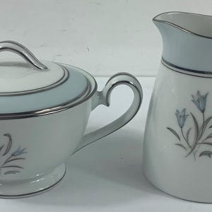 May include: A white ceramic sugar bowl with a lid and a creamer pitcher. Both pieces have a light blue band with silver trim and a floral design. The sugar bowl has two handles and the creamer has one handle.