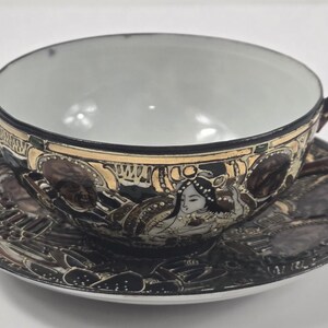 May include: A decorative teacup and saucer set. The cup and saucer are black with gold accents and feature a detailed illustration of a woman. The cup has a dark brown handle.